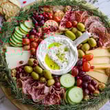 Beautiful evergreen wreath board recipe: creamy dip surrounded by cheese, fruit, and rosemary sprigs.